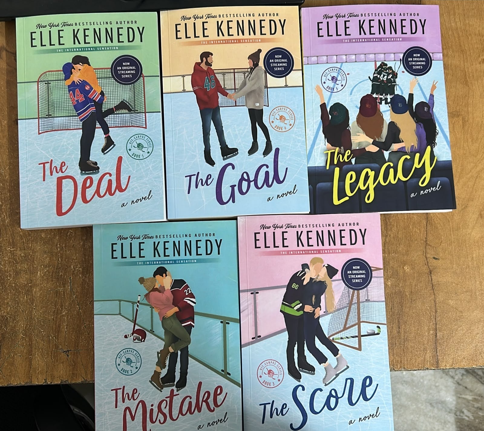 Six hardcover books by Elle Kennedy arranged in two rows on a wooden tabletop. The books are The Mistake, The Deal, The Goal, The Scare, and The Legacy, each with a different pastel-colored cover featuring illustrated characters in sports poses.