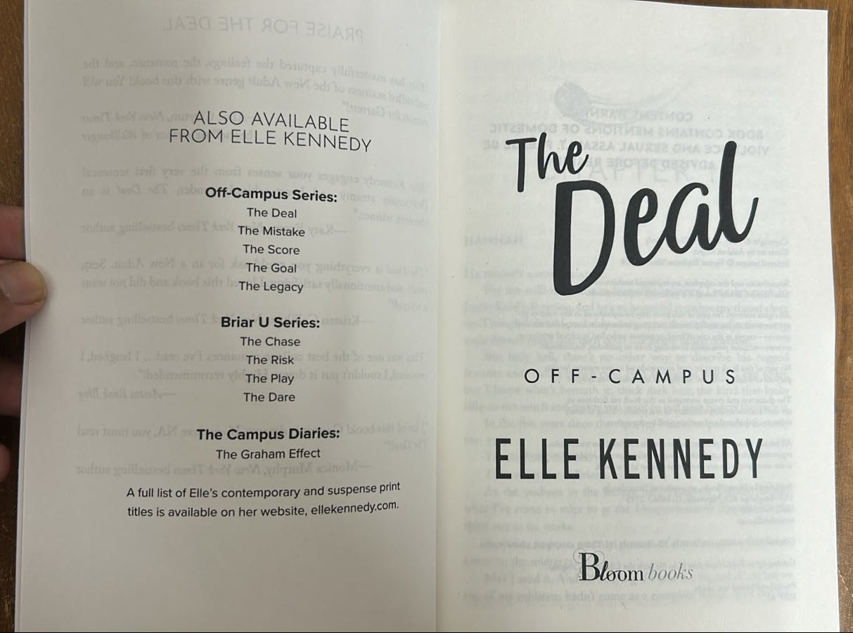 A hand holding an open book or booklet on a wooden surface. The pages show the title 'The Decal' and 'Off Campus' with 'Bloom Books' visible at the bottom.