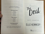 A hand holding an open book or booklet on a wooden surface. The pages show the title 'The Decal' and 'Off Campus' with 'Bloom Books' visible at the bottom.