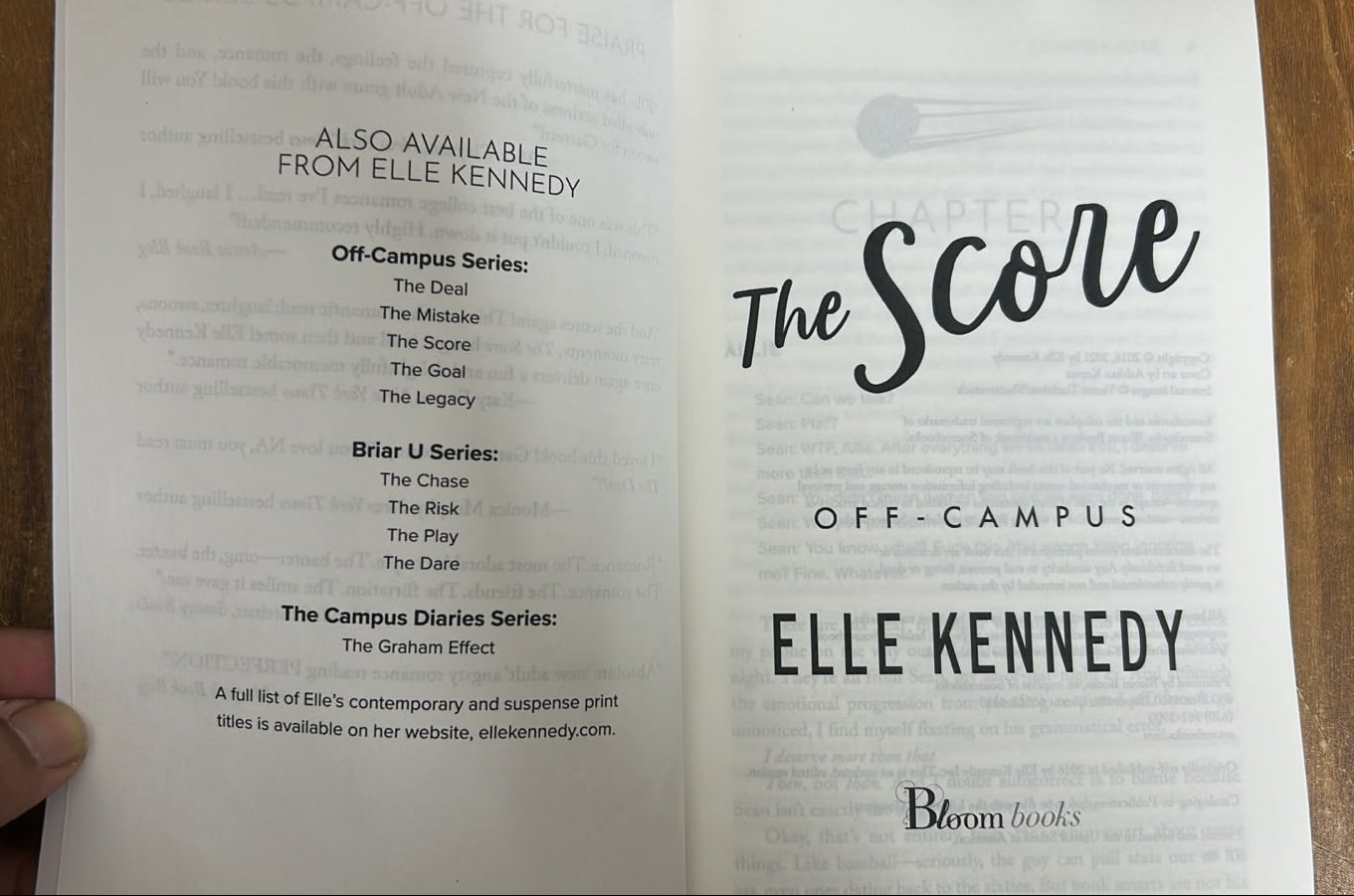 An open book showing the title page for 'The Scene' Off-Campus by Elle Kennedy, photographed on a wooden tabletop with a hand holding the top edge.