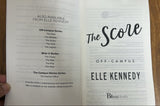 An open book showing the title page for 'The Scene' Off-Campus by Elle Kennedy, photographed on a wooden tabletop with a hand holding the top edge.
