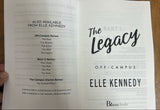 A hand holds an open book or booklet showing printed pages with the title 'The Legacy' and 'ELLE KENNEDY OFF-CAMPUS' visible, photographed on a wooden surface.