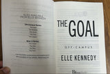 An open book or booklet showing printed text and large title text reading 'THE GOAL' and 'ELLE KENNEDY OFF-CAMPUS' on a wooden surface, with a hand holding one page edge.